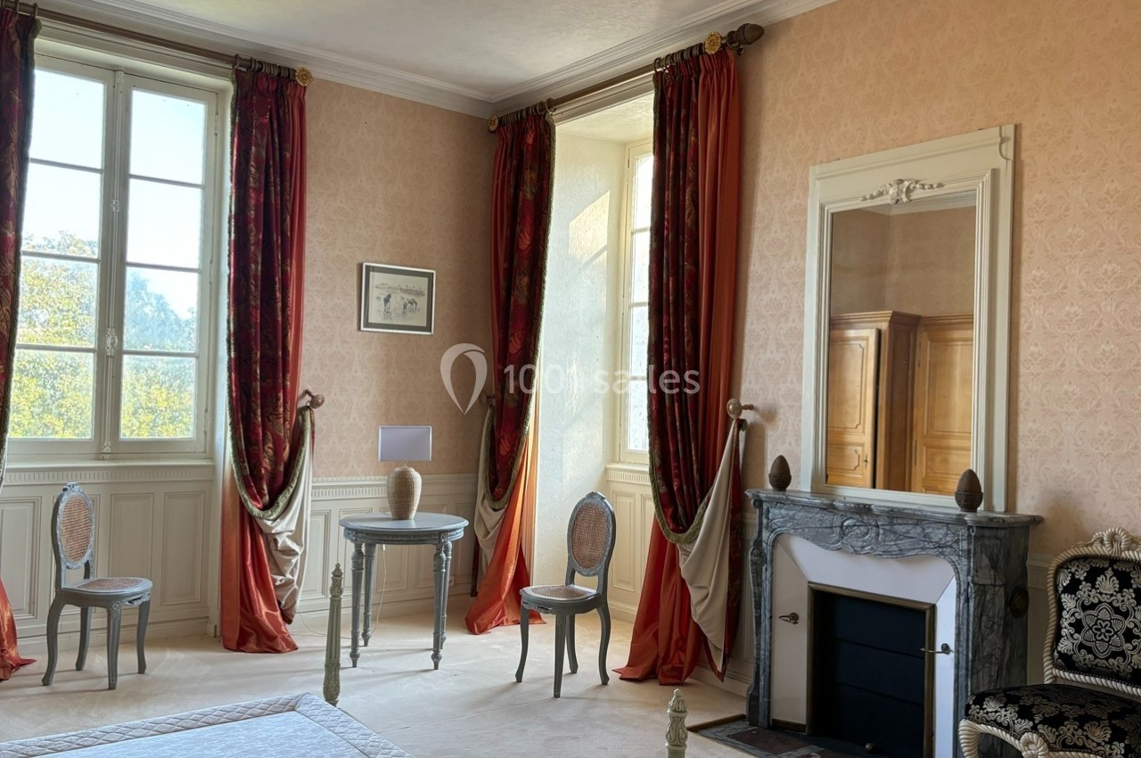 Chambre lumineuse avec murs ornés, cheminée en marbre, rideaux rouges et mobilier classique près de grandes fenêtres.