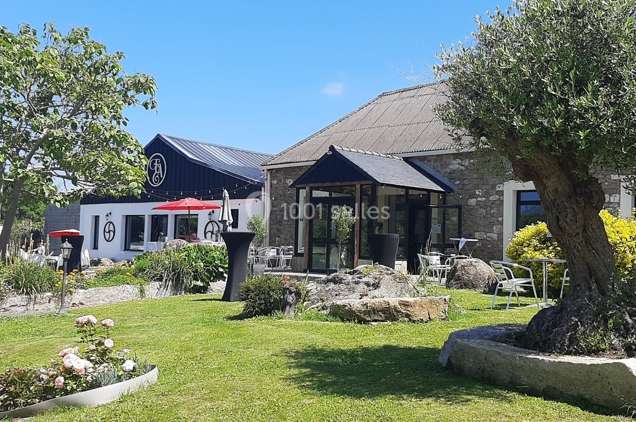 Jardin paysager avec pelouse, fleurs et arbres, devant un bâtiment en pierre et une terrasse avec parasols rouges.