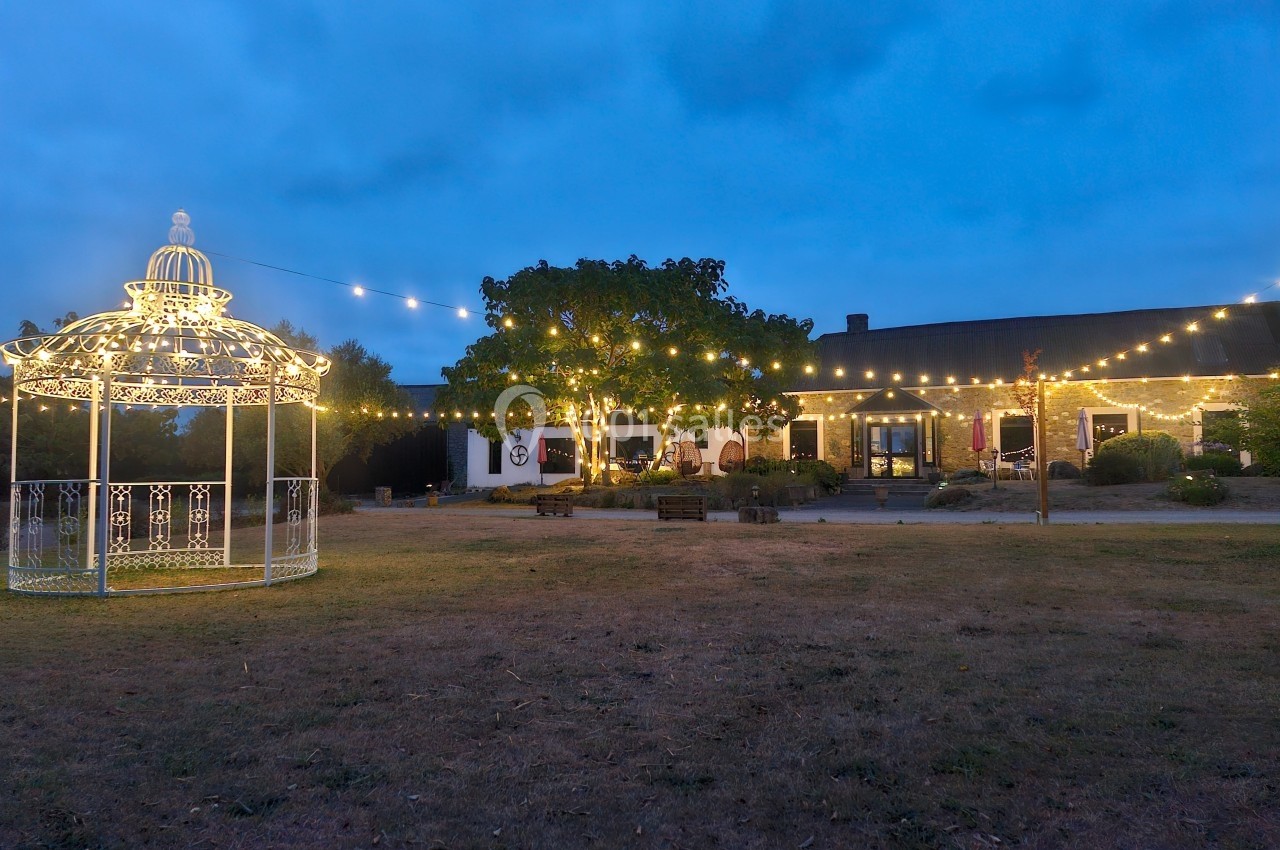 Jardin éclairé au crépuscule avec un kiosque illuminé et des guirlandes lumineuses suspendues devant un bâtiment.
