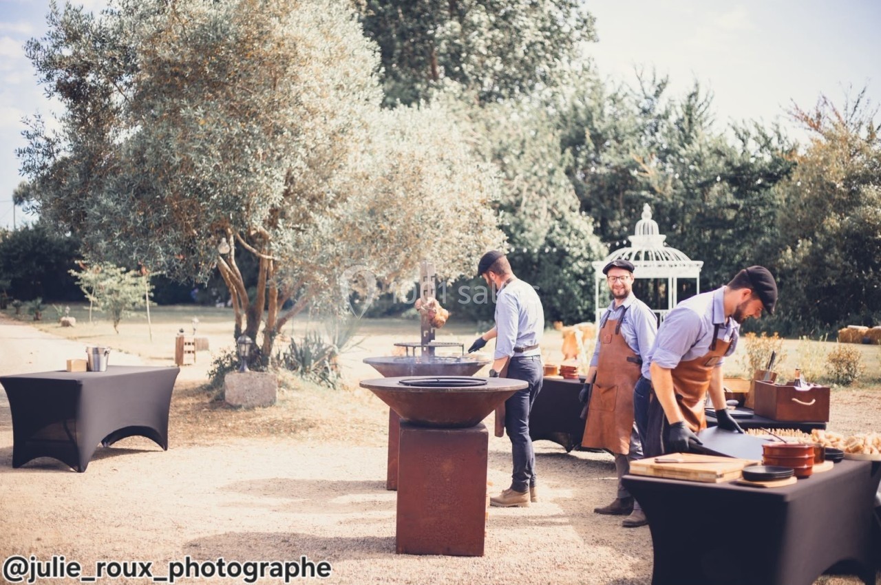 Trois cuisiniers préparent des plats sur des braseros dans un jardin avec des tables et une tonnelle en arrière-plan.