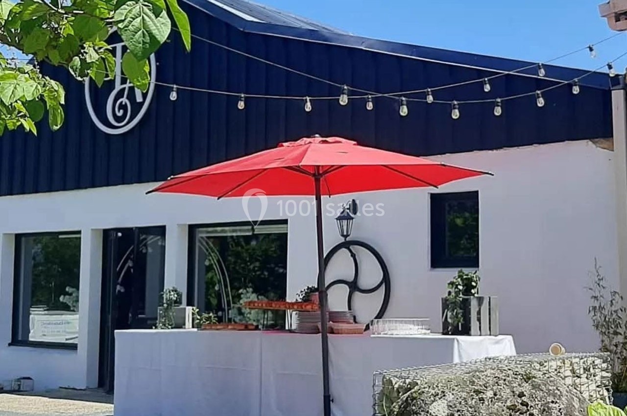 Façade d'un bâtiment moderne avec une table dressée sous un parasol rouge, entourée de décorations lumineuses.