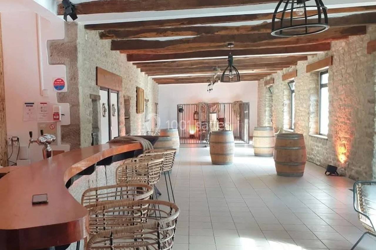Salle lumineuse avec murs en pierre, poutres apparentes, bar en bois, tonneaux décoratifs et chaises en rotin.