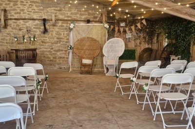 Salle de cérémonie rustique avec chaises blanches alignées, décor floral et fauteuils en osier devant un mur en pierre.