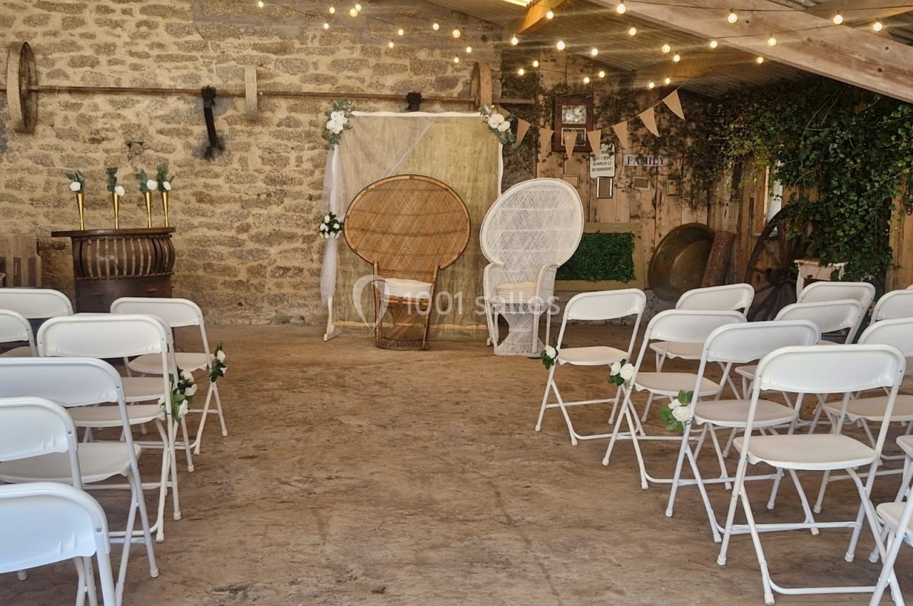Salle de cérémonie rustique avec chaises blanches alignées, décor floral et fauteuils en osier devant un mur en pierre.