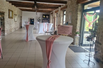 Salle de cérémonie rustique avec chaises blanches alignées, décor floral et fauteuils en osier devant un mur en pierre.