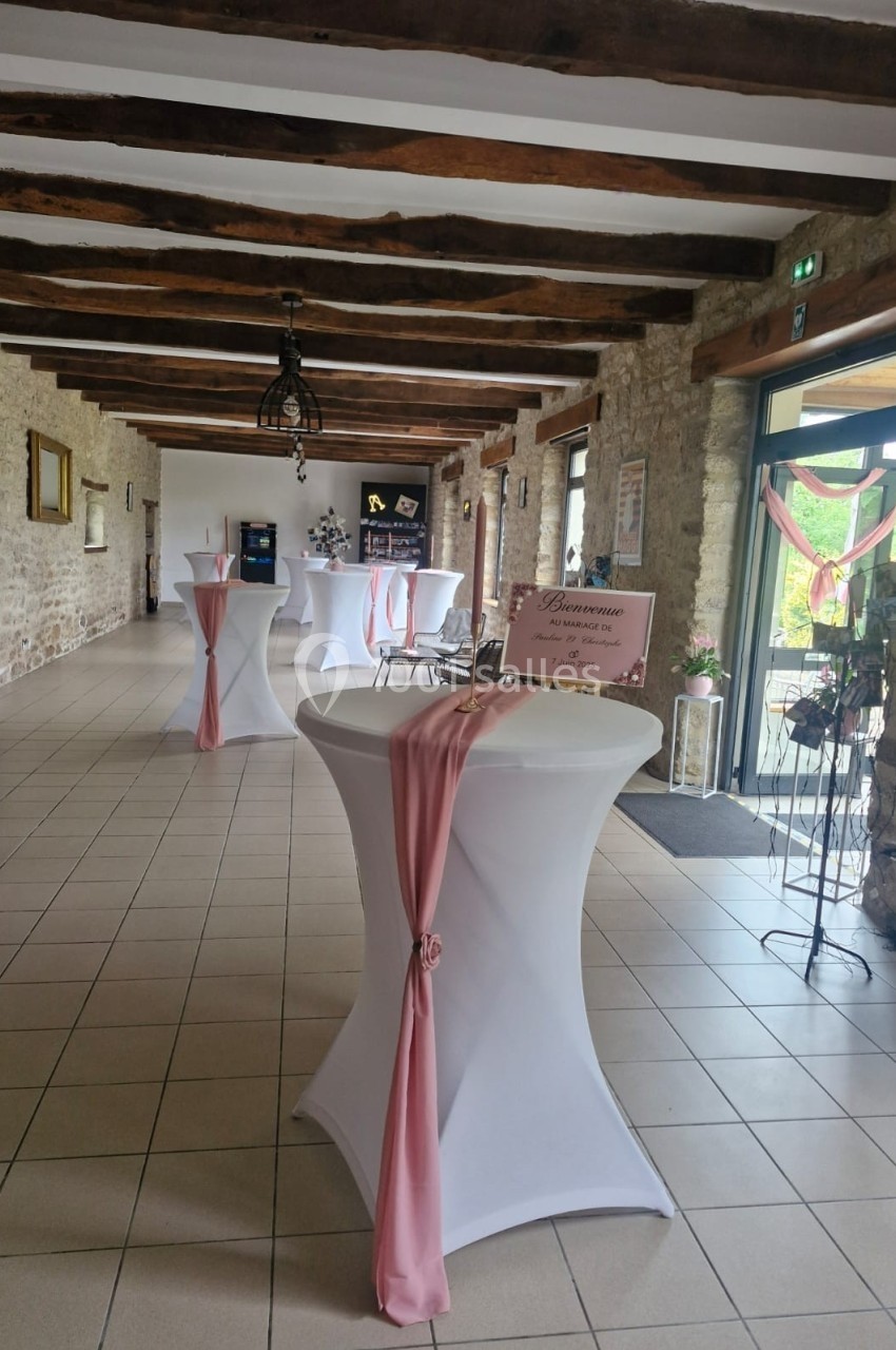 Salle de réception avec tables hautes drapées de tissu rose, poutres apparentes et lumière naturelle.