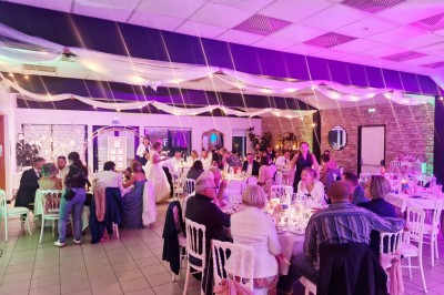 Salle de cérémonie rustique avec chaises blanches alignées, décor floral et fauteuils en osier devant un mur en pierre.