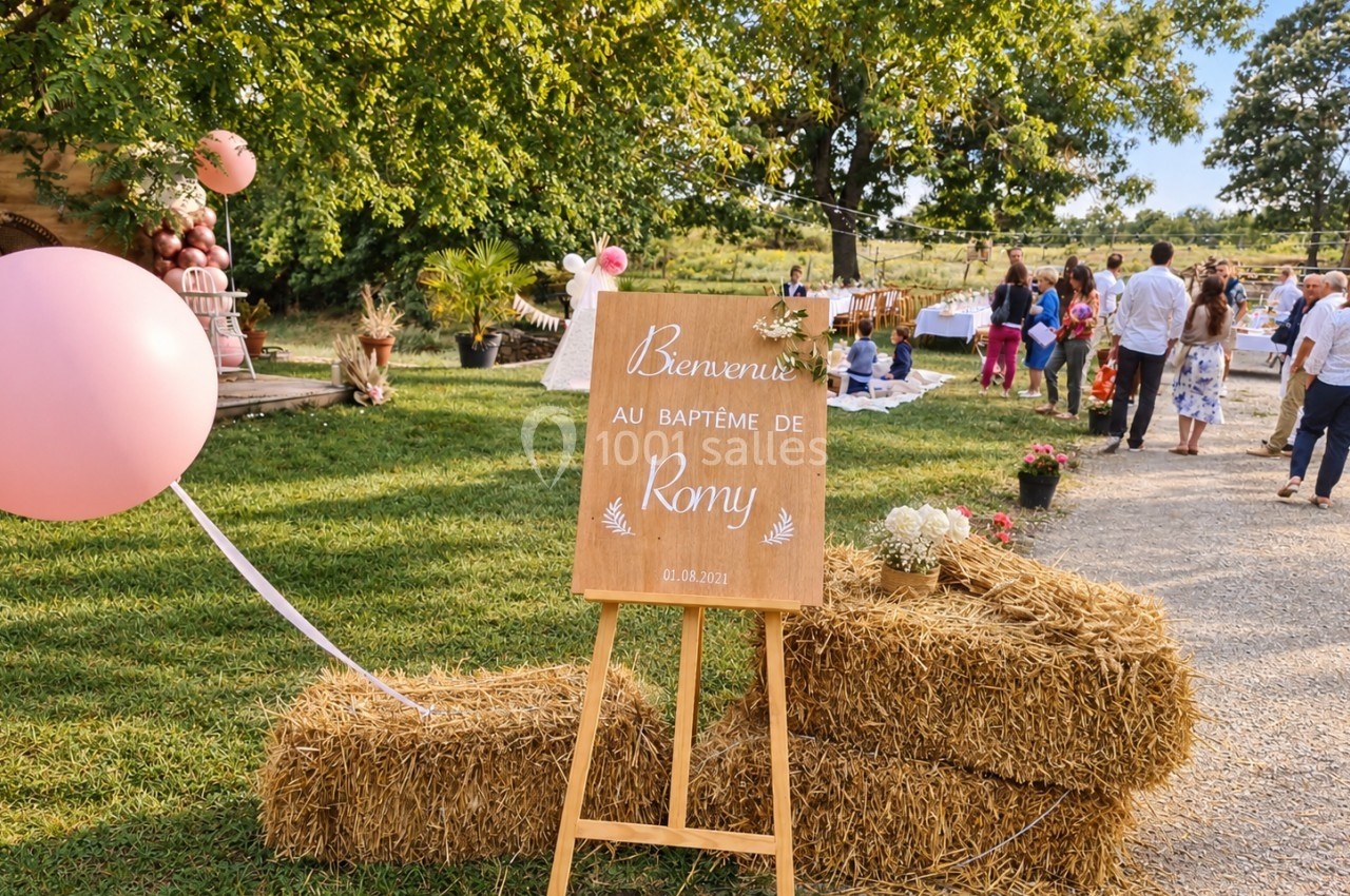 Panneau en bois indiquant ’Bienvenue au baptême de Romy’ posé sur des bottes de paille dans un cadre champêtre.