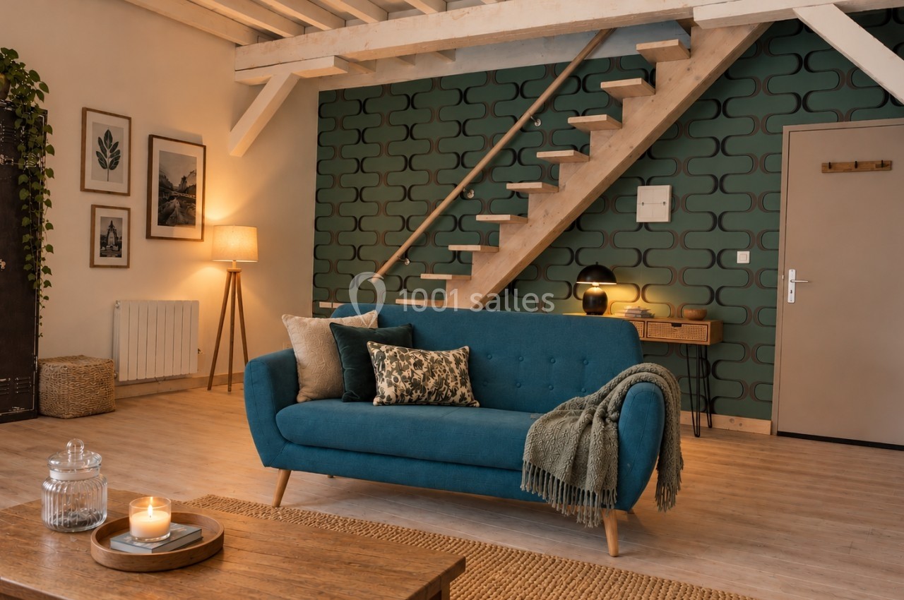 Salon cosy avec canapé bleu, mur vert à motifs, escalier en bois, table basse et éclairage chaleureux.