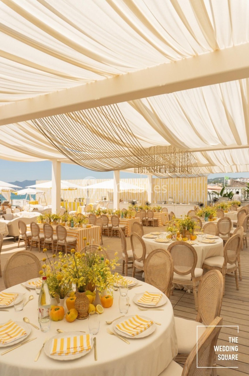 Salle de réception en plein air avec tables décorées de fleurs jaunes et citrons, sous une pergola blanche.