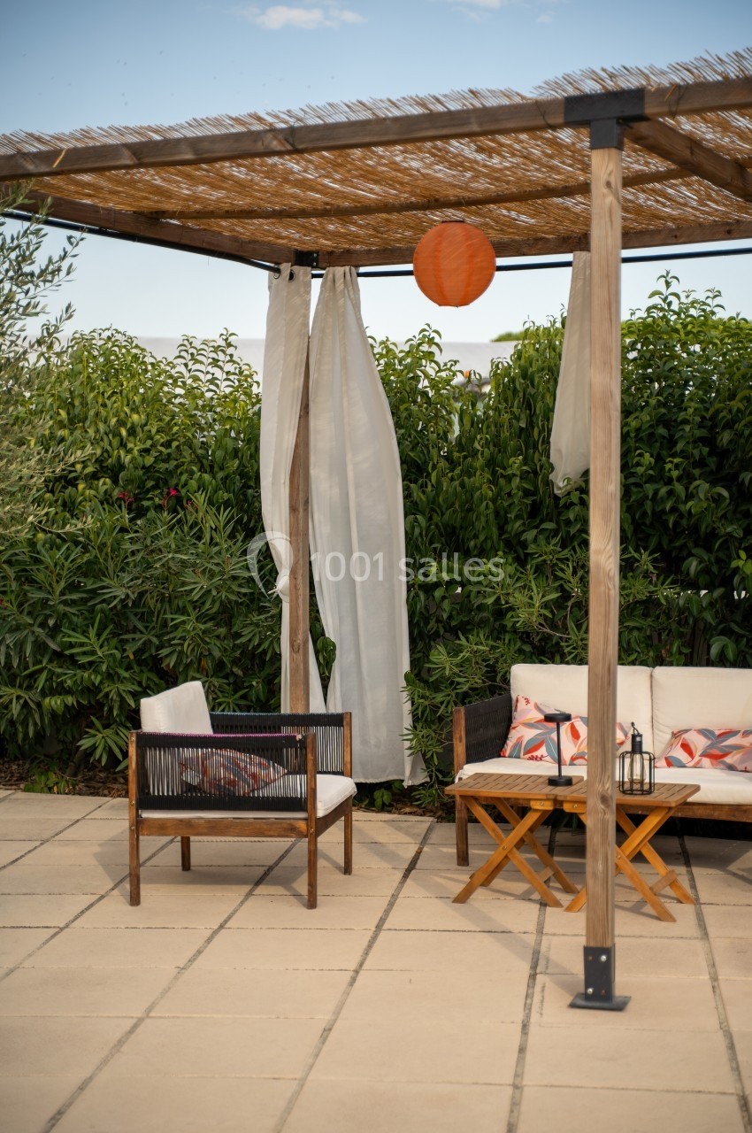 Pergola en bois avec rideaux blancs, fauteuils et table basse sur une terrasse carrelée, entourée de végétation.