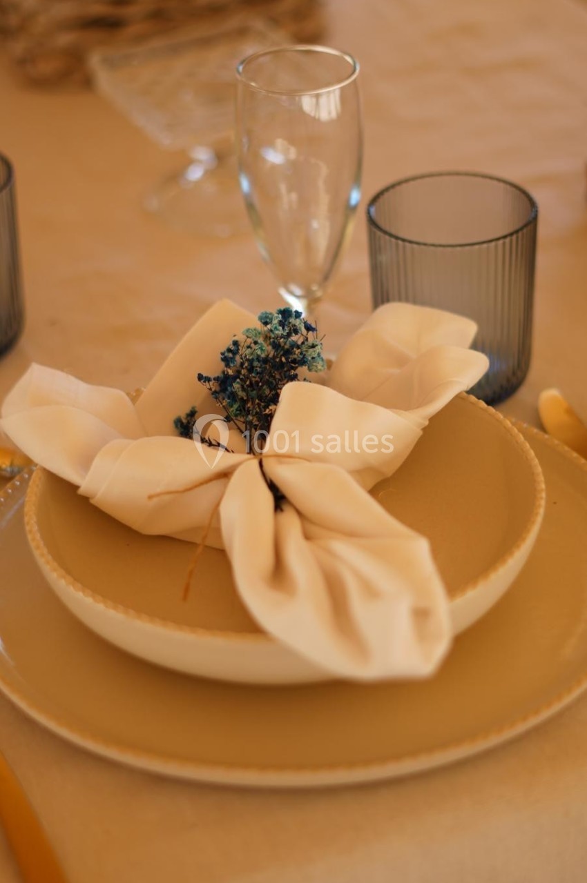 Serviette nouée avec des fleurs séchées sur une assiette beige, entourée de verres et couverts dorés.