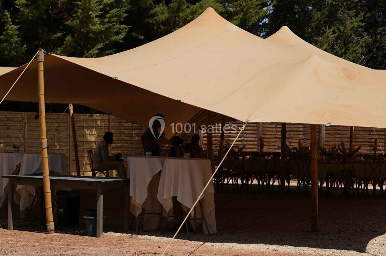 Tente beige installée en extérieur, abritant des tables hautes et des chaises, entourée de clôtures en bois.