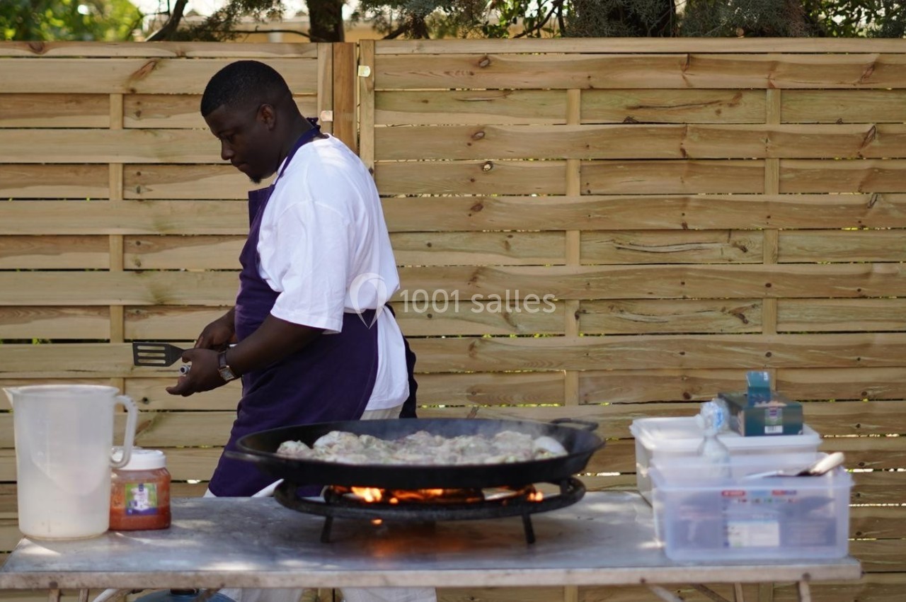 Un cuisinier en tablier violet prépare des aliments sur une grande poêle chauffée, devant une clôture en bois.