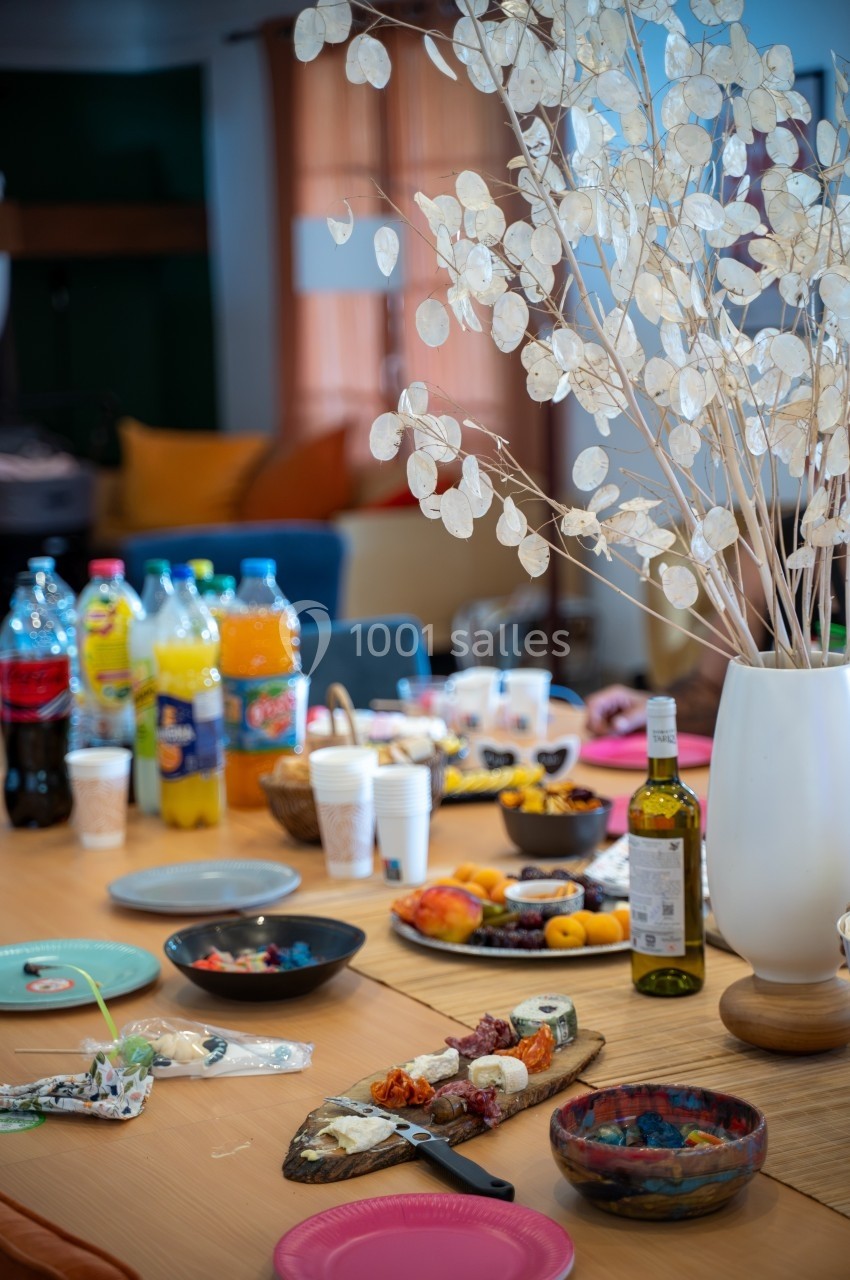 Table garnie de boissons, plats variés et décorée d'un vase blanc avec des branches, dans un cadre intérieur chaleureux.