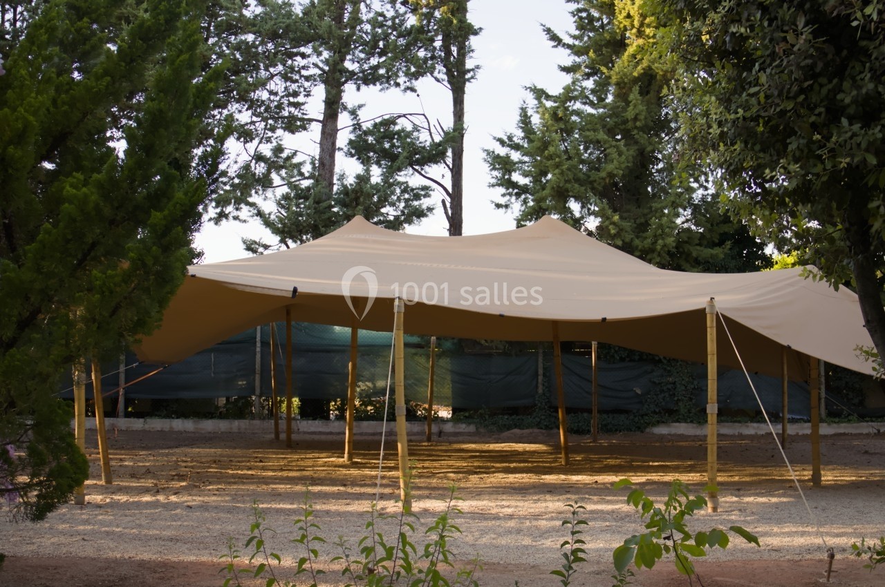 Grande tente beige montée sur des poteaux en bois, installée dans un espace extérieur entouré d'arbres.
