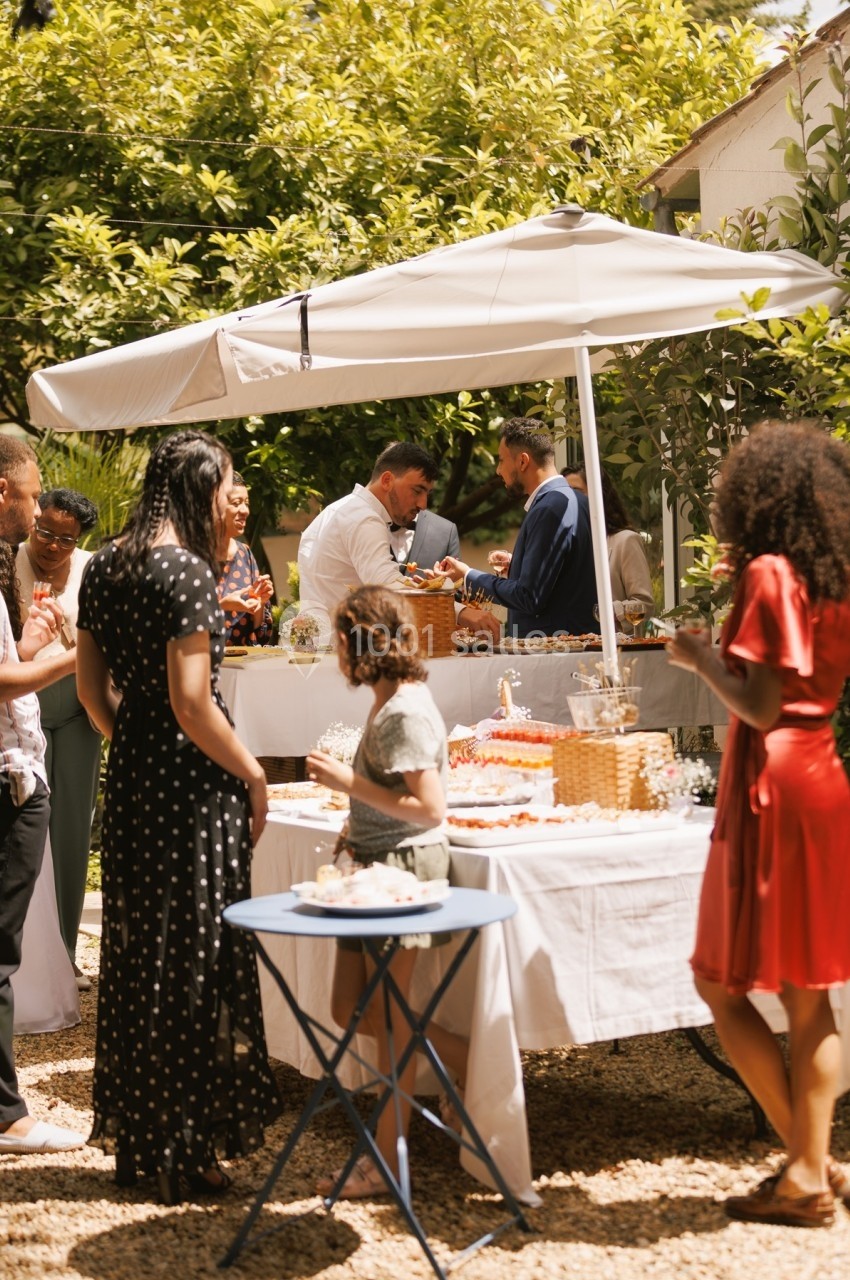 Groupe de personnes réunies autour d'une table avec buffet en extérieur, sous un parasol, par une journée ensoleillée.