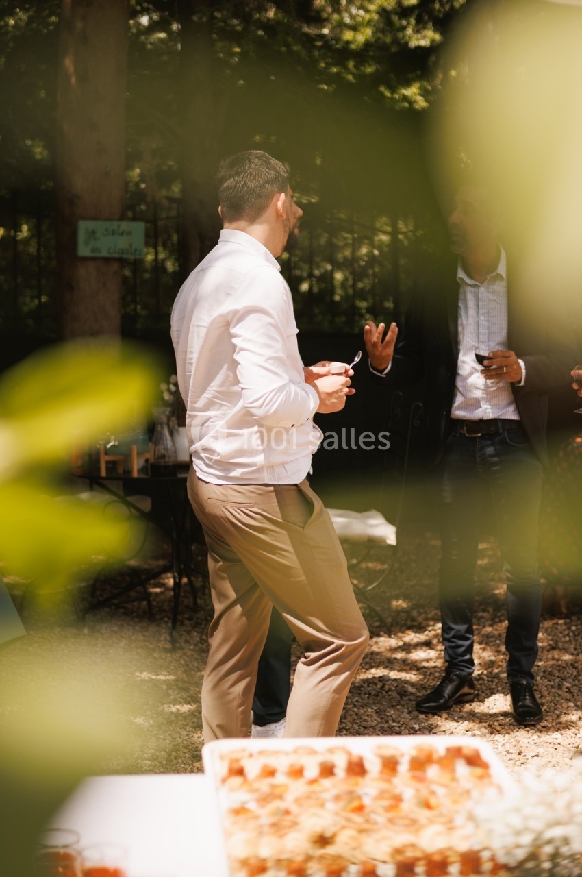 Un homme en chemise blanche discute avec d'autres personnes dans un jardin lors d'un événement en plein air.
