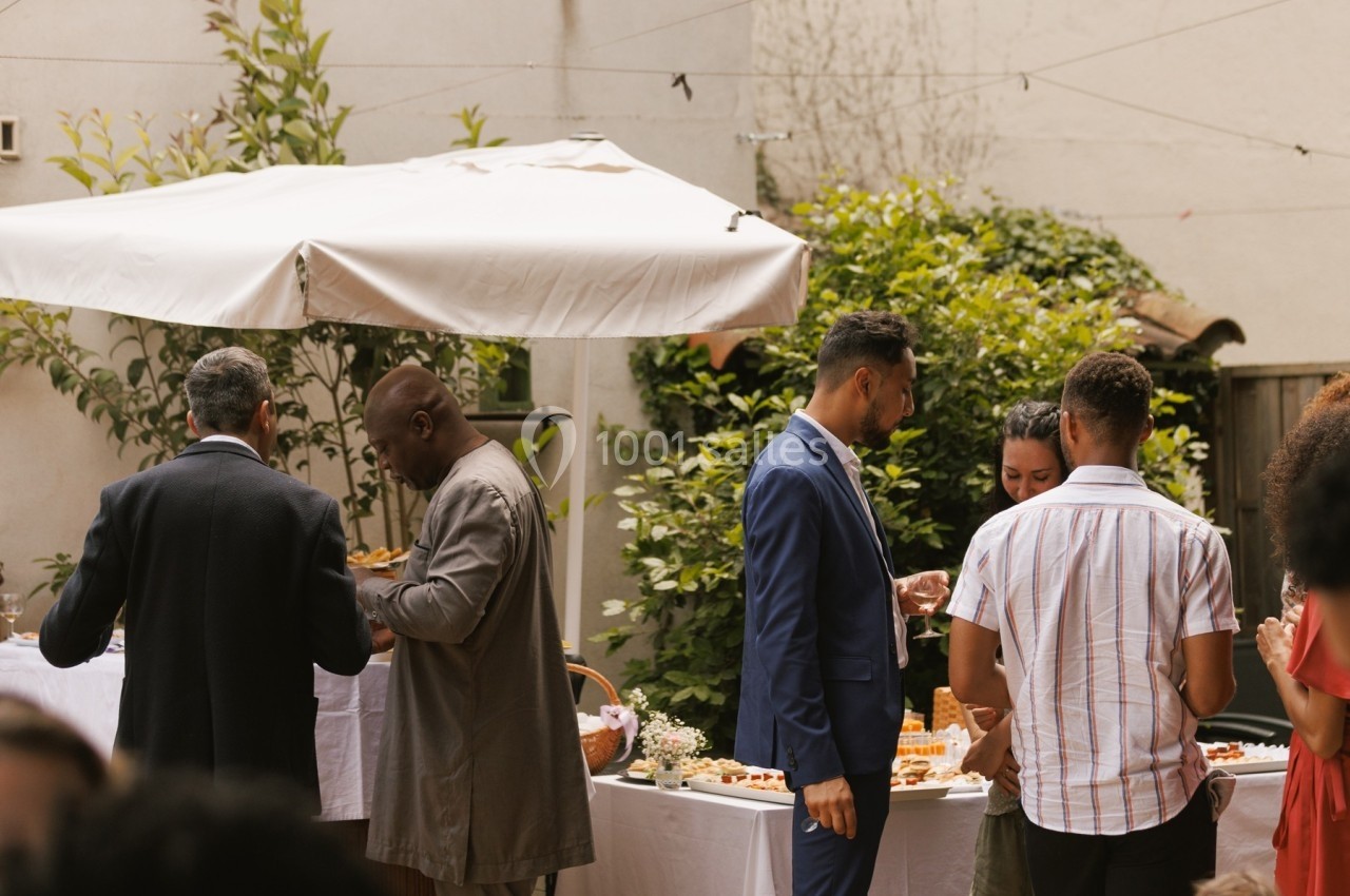Des personnes discutent et se servent à un buffet en extérieur, sous un parasol, dans un cadre verdoyant.