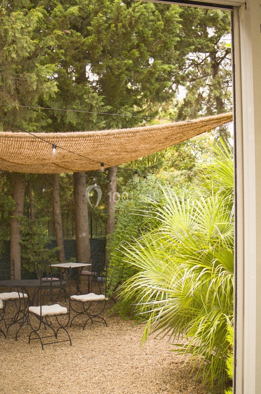 Vue d'un jardin avec des chaises et une table sur gravier, ombragé par une toile beige et entouré de végétation dense.