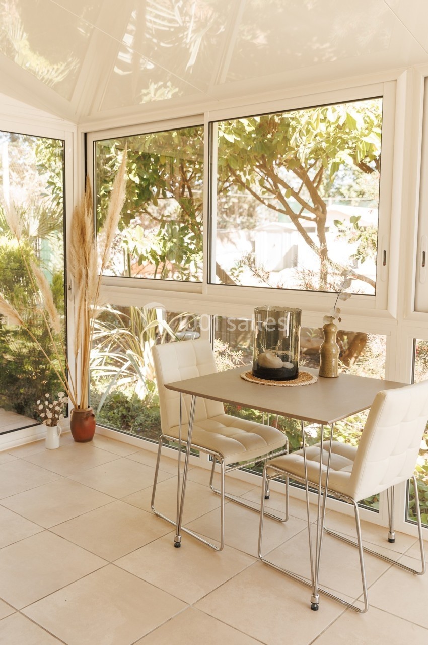 Table et chaises blanches dans une véranda lumineuse avec vue sur un jardin verdoyant.