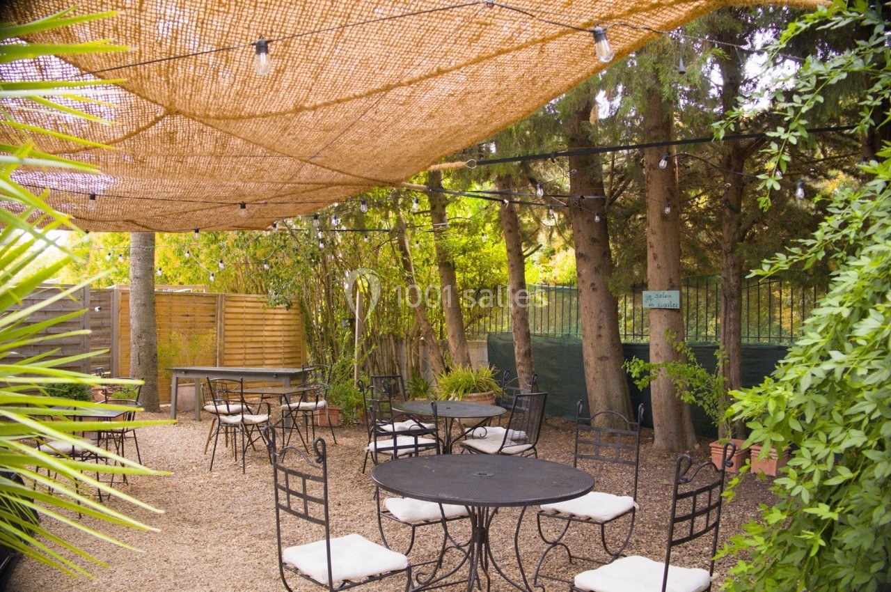 Terrasse extérieure ombragée avec tables et chaises en métal, entourée de végétation et éclairée par des guirlandes…