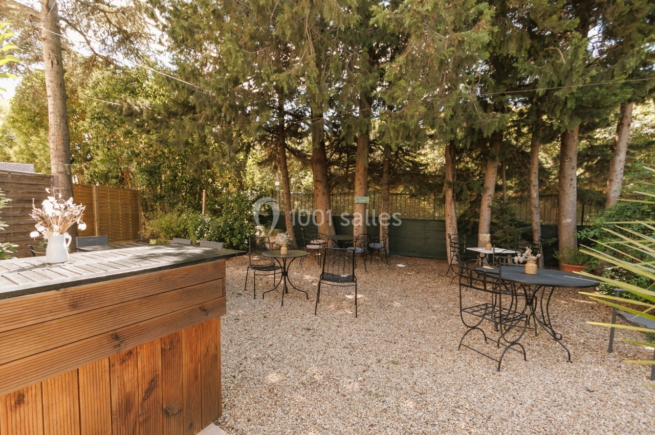 Cour extérieure avec tables et chaises en métal sur un sol en gravier, entourée d'arbres et de verdure.