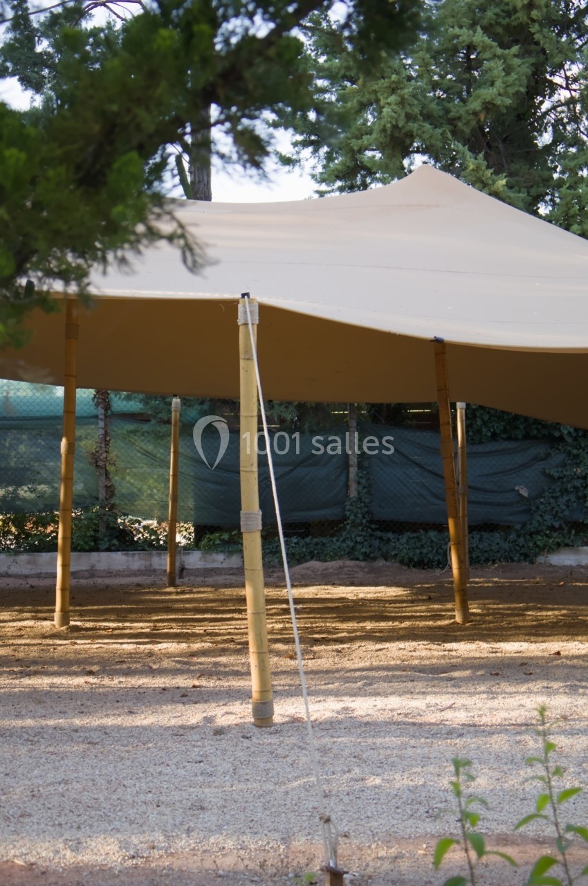 Tente beige montée sur des poteaux en bambou, installée en extérieur avec des arbres et des clôtures en arrière-plan.