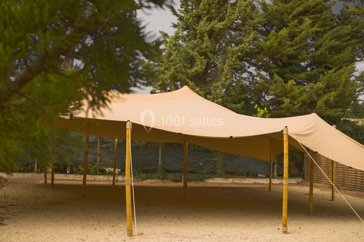 Tente beige en toile tendue sur des poteaux en bambou, installée sur un sol en gravier entouré d'arbres.