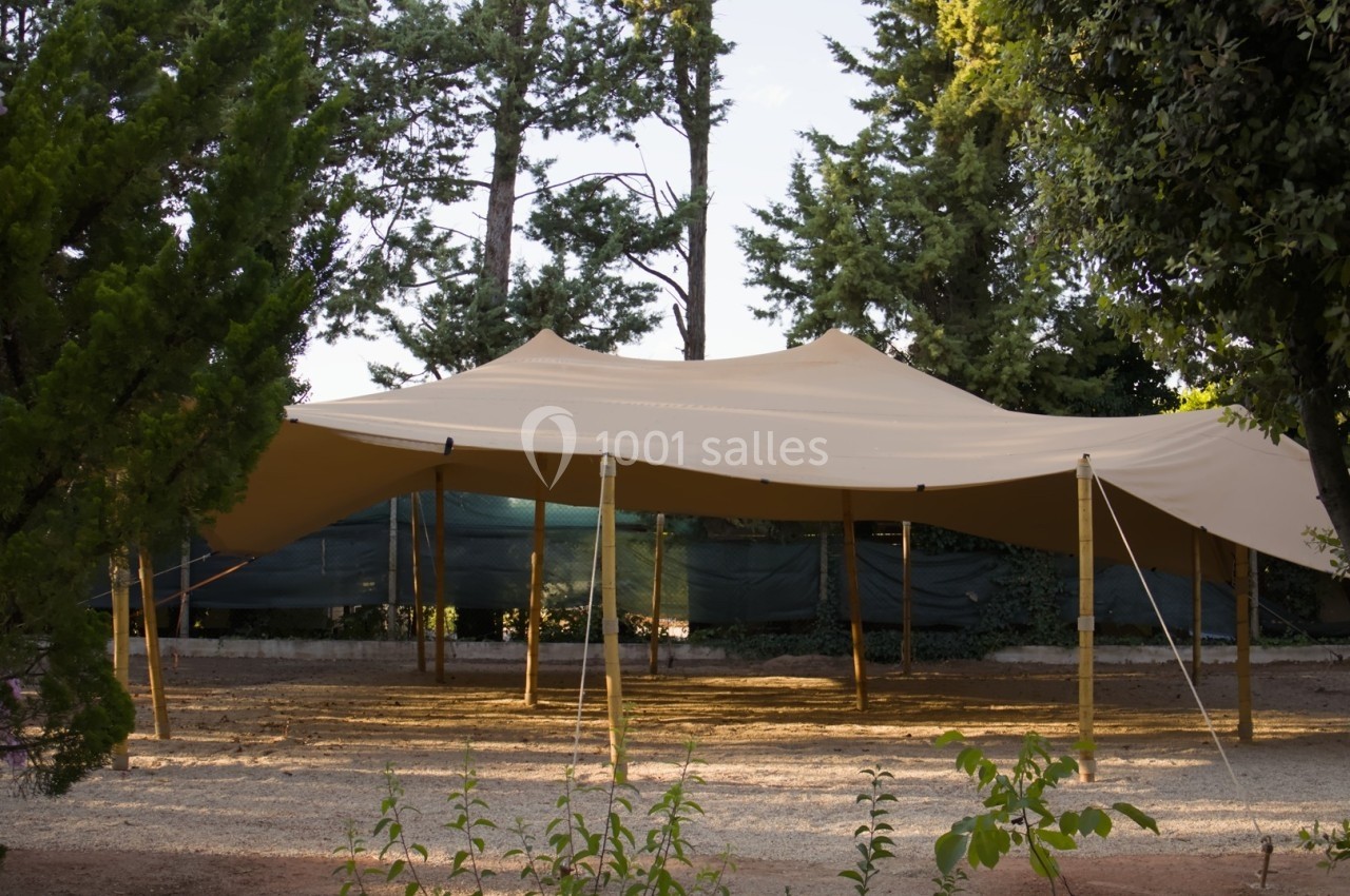 Tente beige tendue sur des poteaux en bois, installée dans un espace extérieur entouré d'arbres et de végétation.