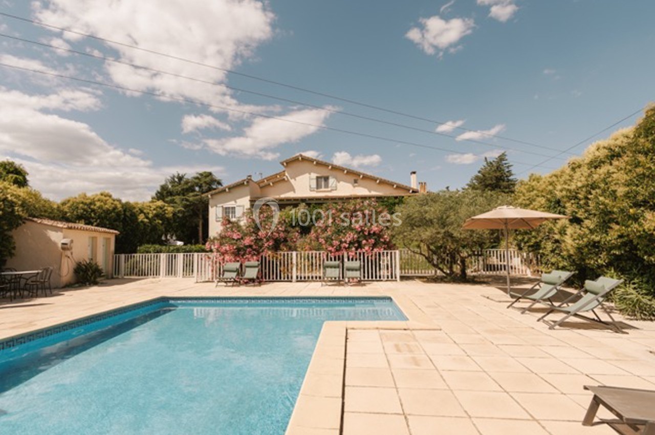 Maison avec jardin, piscine, chaises longues et parasol, entourée de végétation sous un ciel partiellement nuageux.