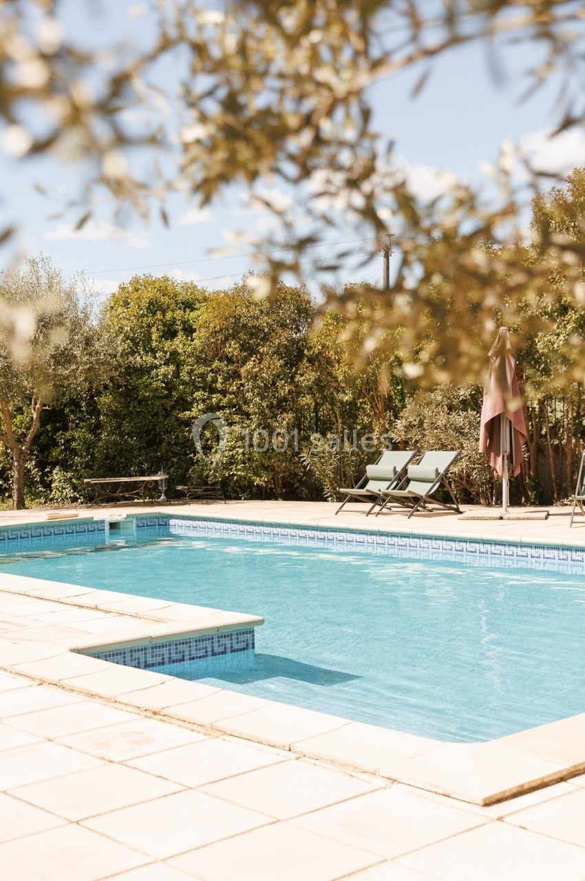 Piscine extérieure entourée de végétation, avec deux chaises longues et un parasol sur une terrasse ensoleillée.