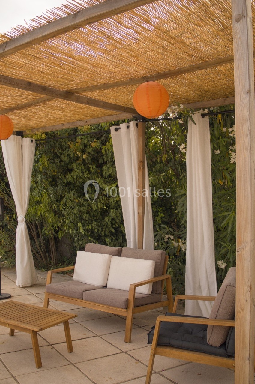 Pergola en bois avec toit en roseaux, rideaux blancs, mobilier de jardin en bois et coussins, entourée de végétation.