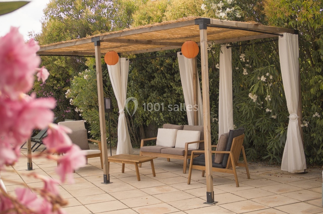 Pergola en bois avec rideaux blancs, mobilier de jardin et lanternes orange, entourée de végétation fleurie.