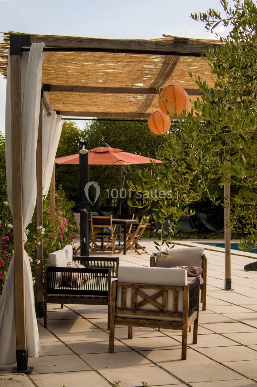 Salon de jardin sous une pergola en bois avec rideaux, près d'une piscine et entouré de végétation.