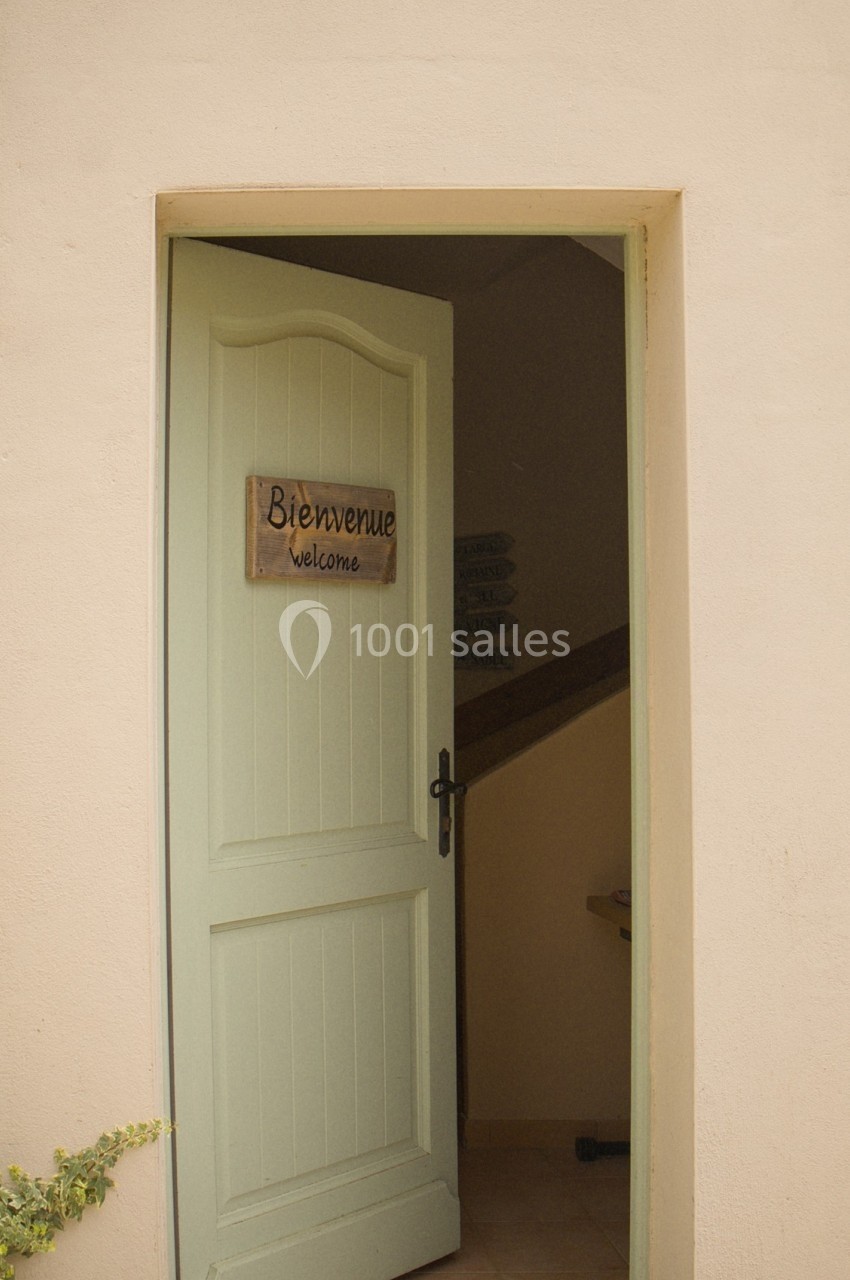 Porte d'entrée beige entrouverte avec un panneau ’Bienvenue’ en français et en anglais, donnant sur un intérieur sombre.