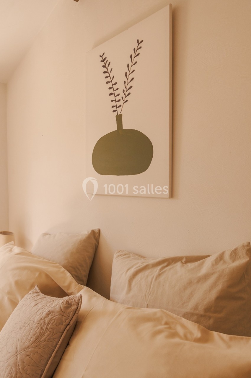Tableau minimaliste avec un vase vert et des branches, accroché au-dessus d'un lit aux draps beiges.