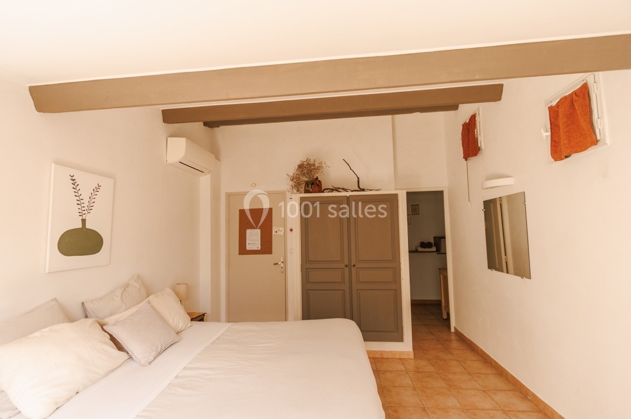 Chambre lumineuse avec lit double, armoire grise, murs blancs, poutres apparentes et décoration minimaliste.