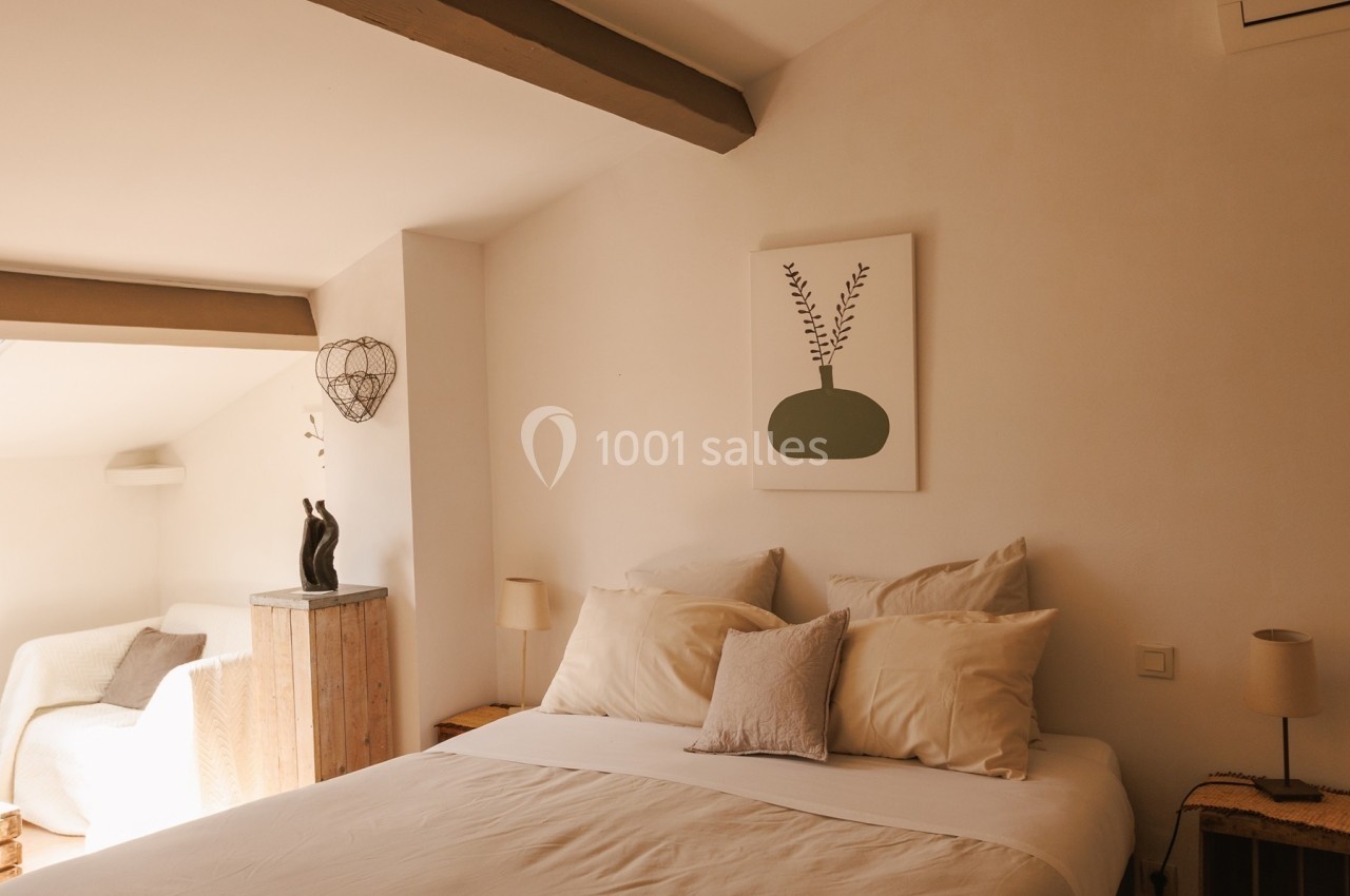 Chambre lumineuse avec lit double, coussins clairs, tableau minimaliste au mur et fauteuil blanc près d'une fenêtre.