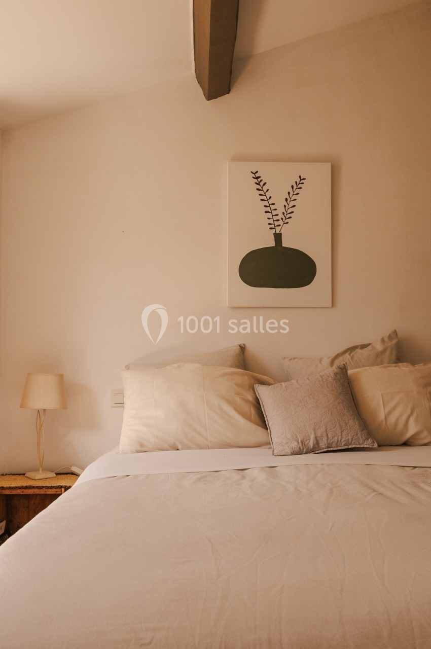 Lit double avec draps beiges, coussins assortis et tableau minimaliste représentant un vase sur le mur.