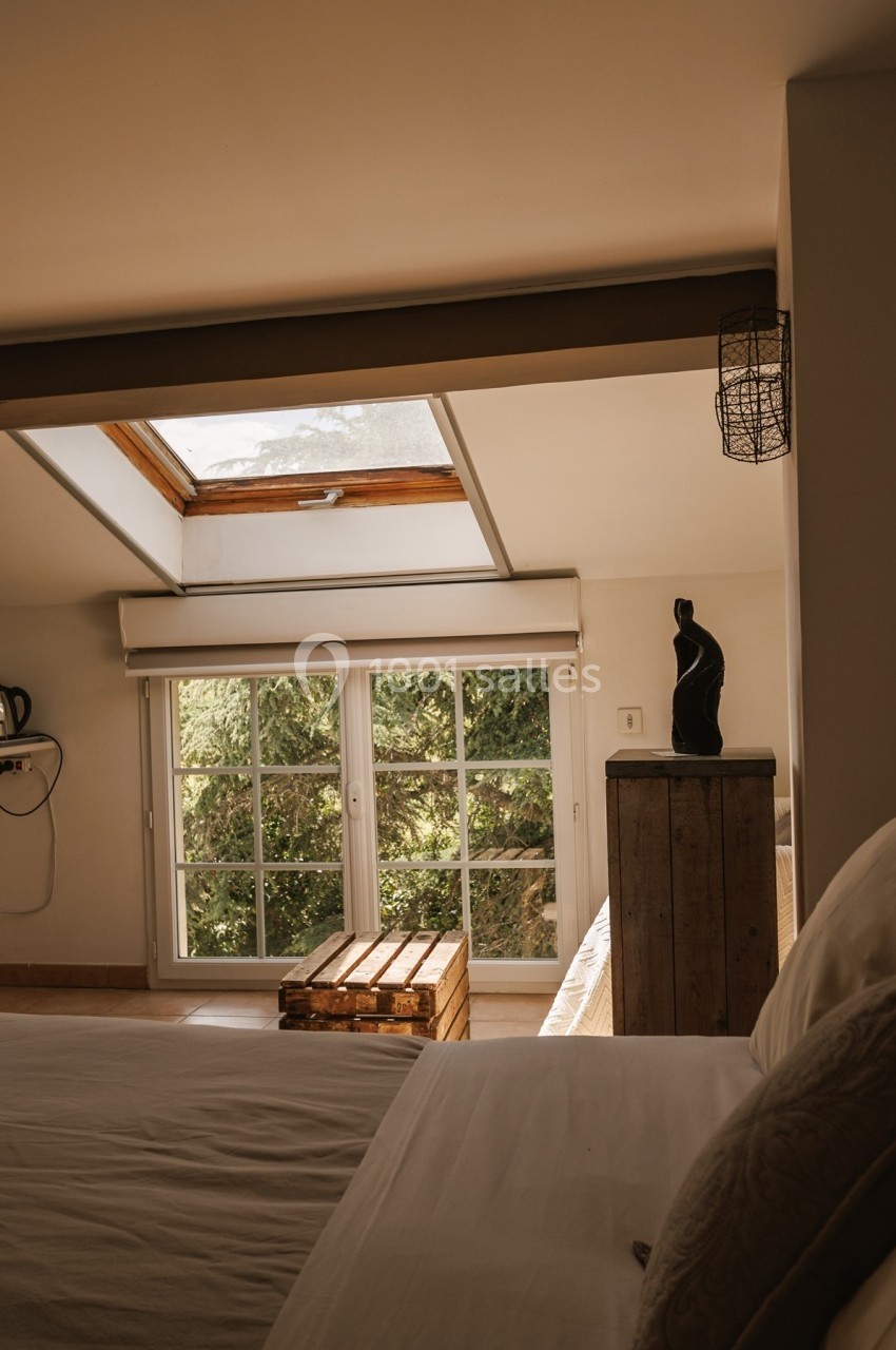 Chambre lumineuse avec lit, fenêtre à battants donnant sur des arbres et puits de lumière au plafond.
