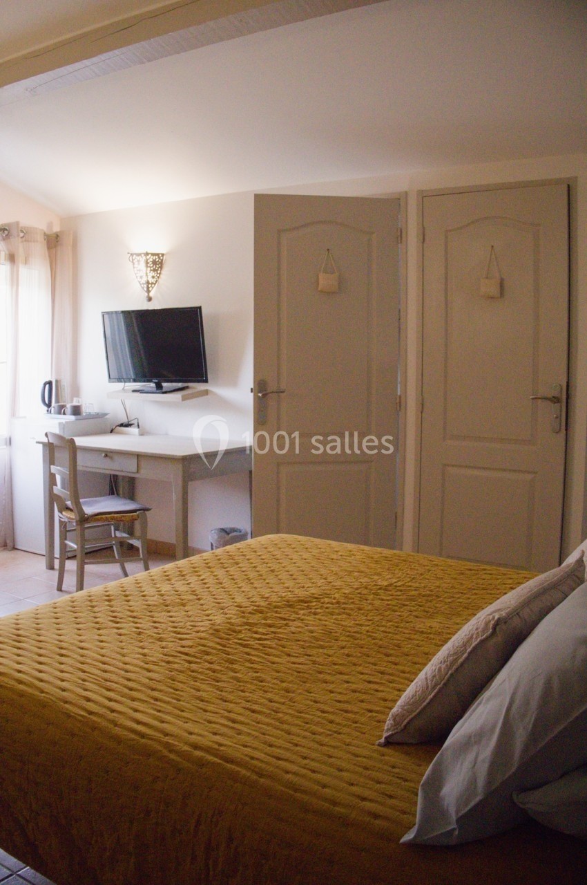Chambre lumineuse avec lit à couverture jaune, bureau, chaise, télévision murale et deux portes blanches.