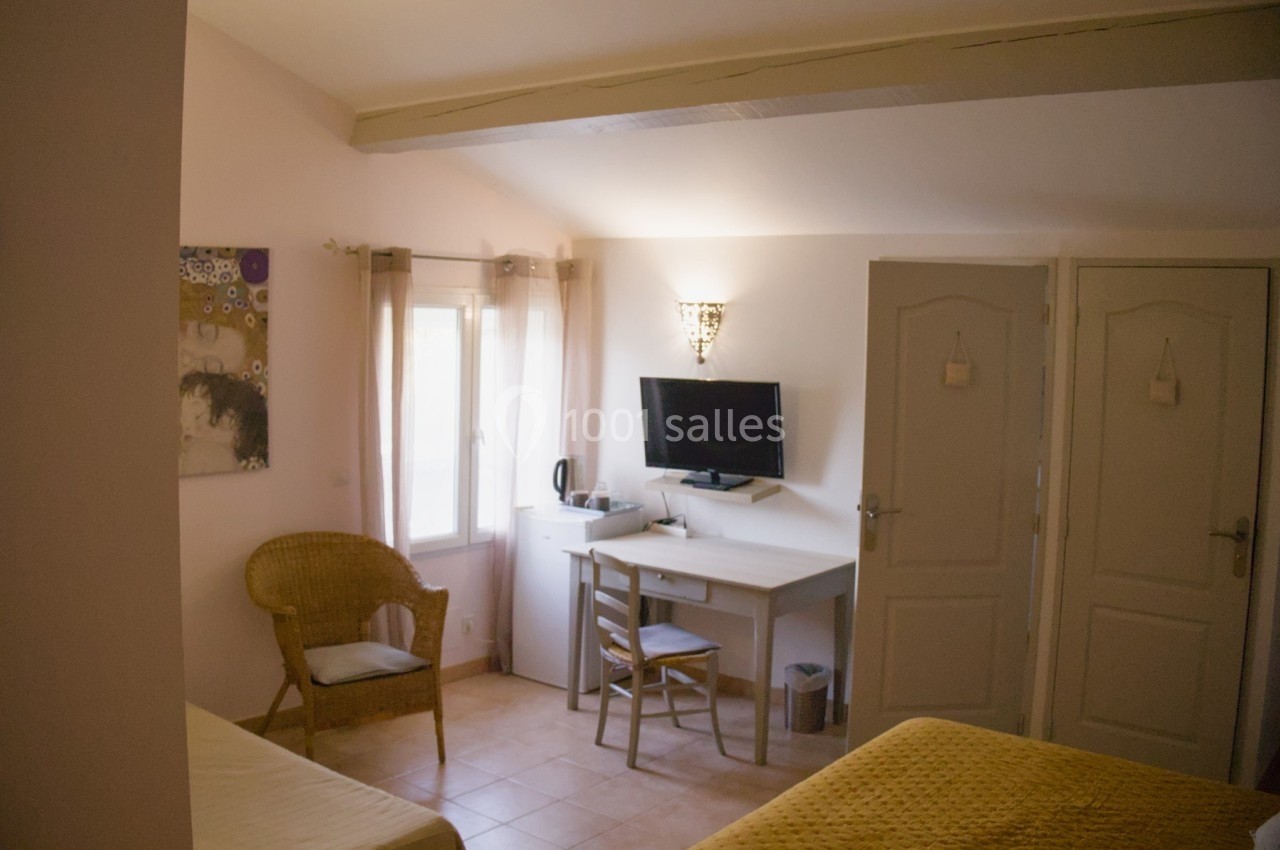 Chambre lumineuse avec bureau, chaise, télévision, lit, fauteuil en osier et fenêtre avec rideaux clairs.