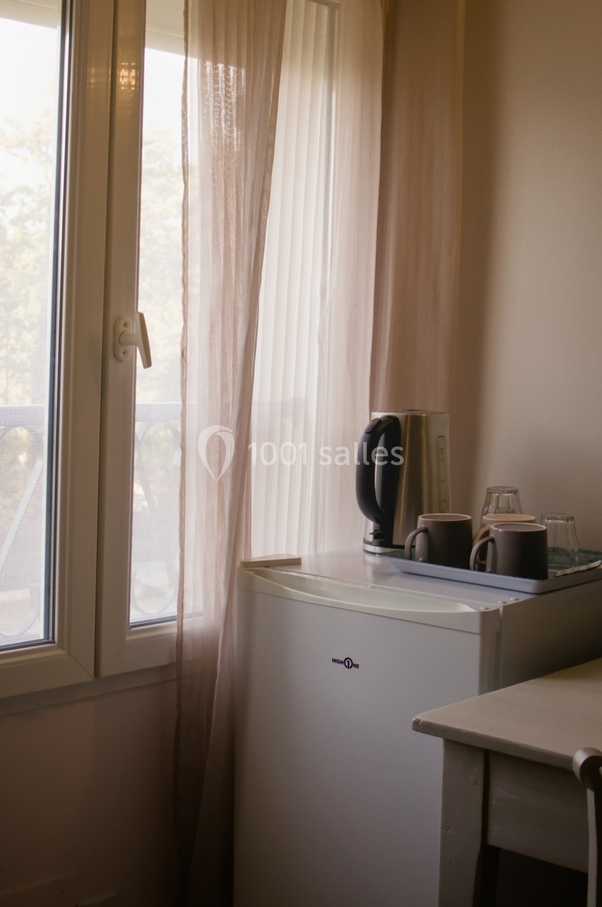 Petite kitchenette avec une bouilloire, des tasses et un mini-réfrigérateur près d'une fenêtre avec rideaux translucides.