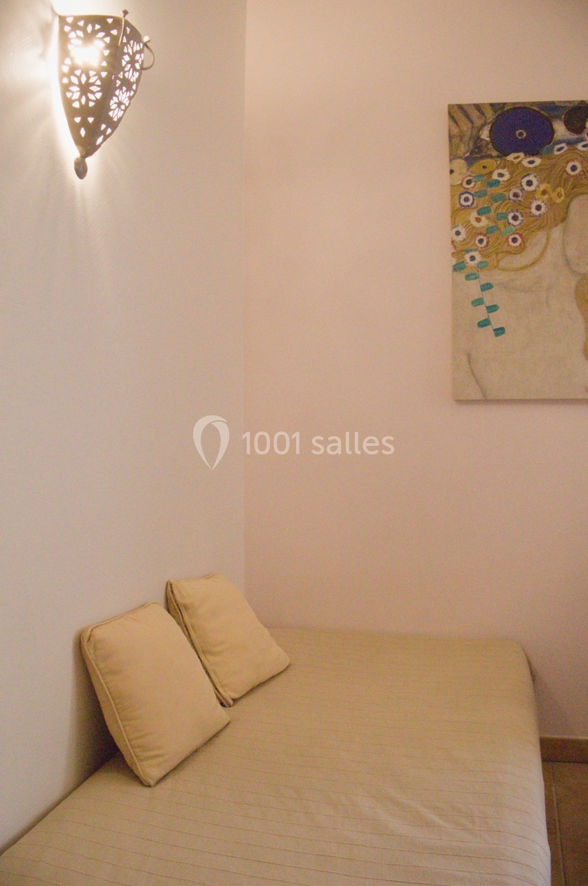 Lit simple avec deux coussins beiges, éclairé par une applique murale décorative et orné d'un tableau coloré.