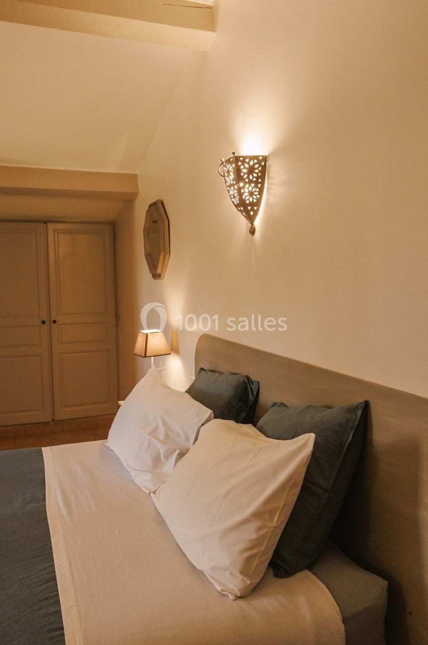 Lit double avec draps blancs et coussins verts, éclairé par une applique murale décorative dans une chambre beige.