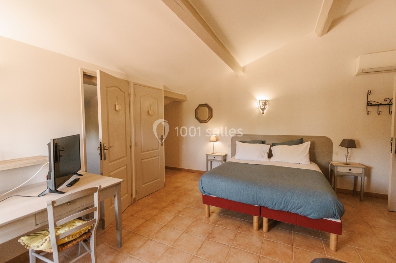 Chambre lumineuse avec lit double, bureau équipé d'un écran, murs blancs et sol en carrelage beige.