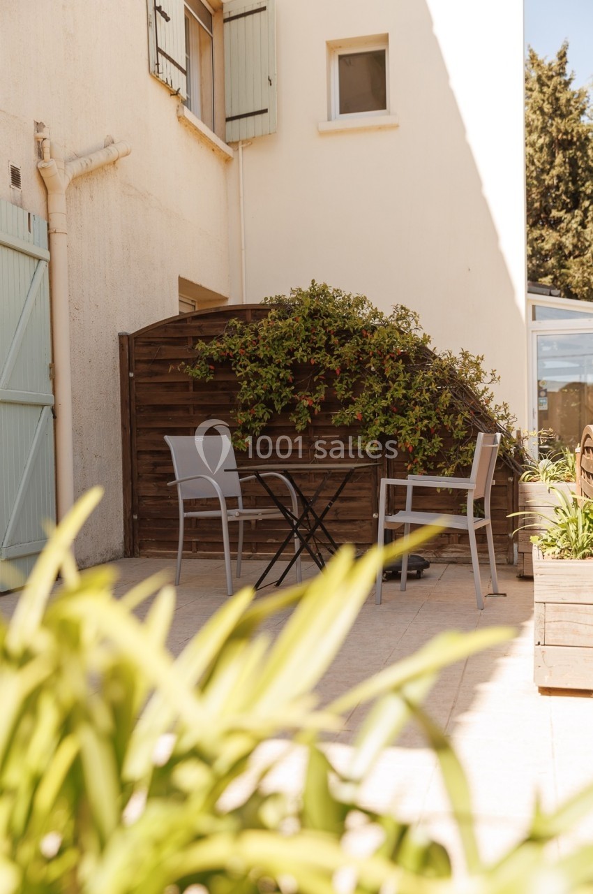 Petite terrasse ensoleillée avec table et chaises, entourée de plantes et adossée à un mur avec volets verts.