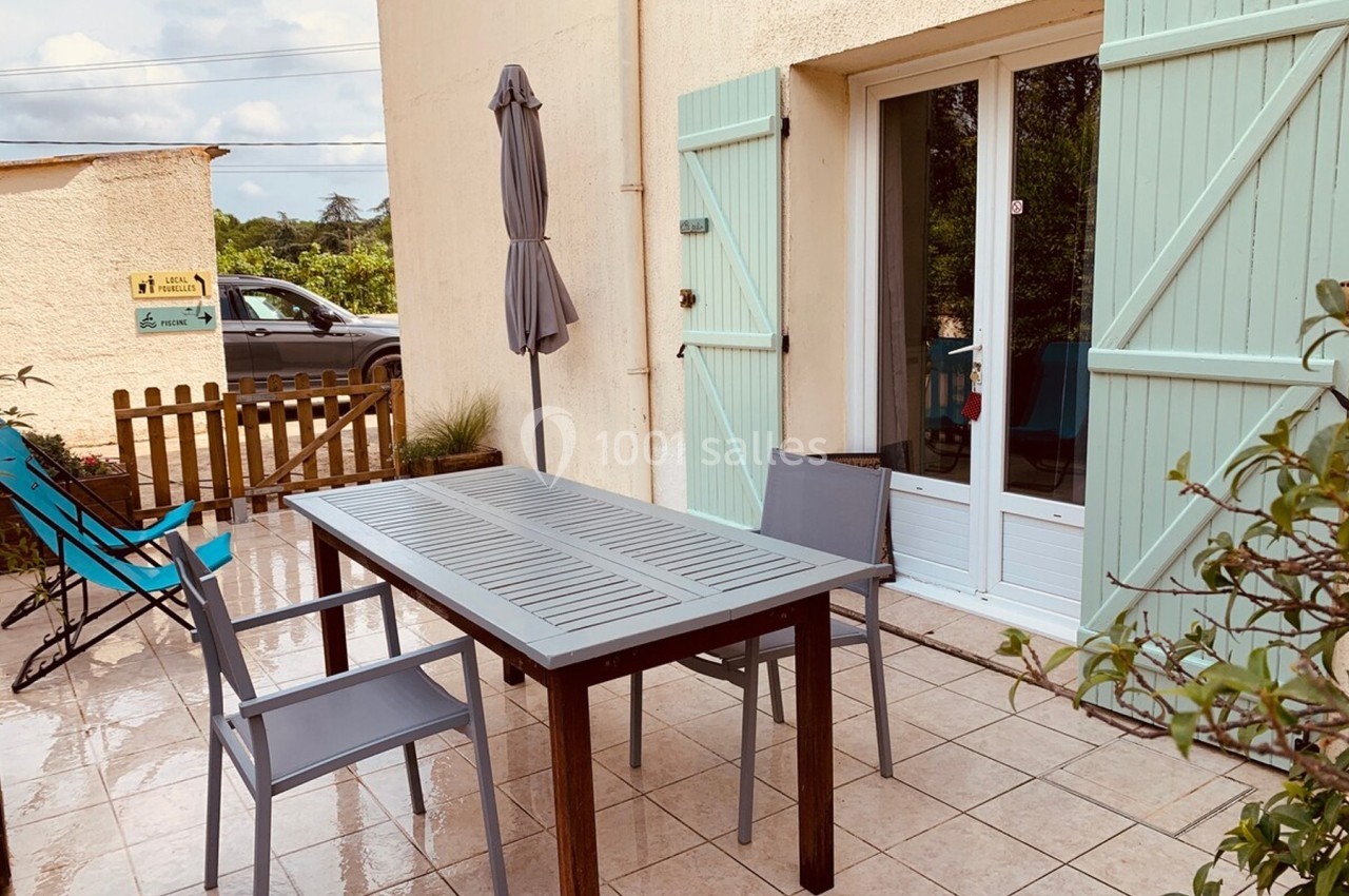 Terrasse carrelée avec table, chaises, parasol fermé et volets verts, donnant sur une porte-fenêtre.