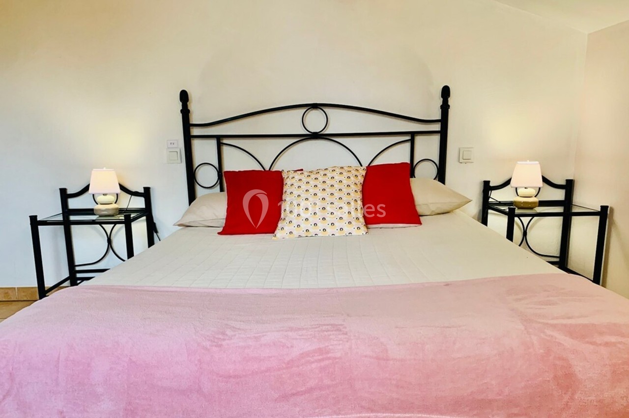 Lit double avec structure en métal noir, draps blancs, plaid rose, coussins rouges et tables de chevet avec lampes.